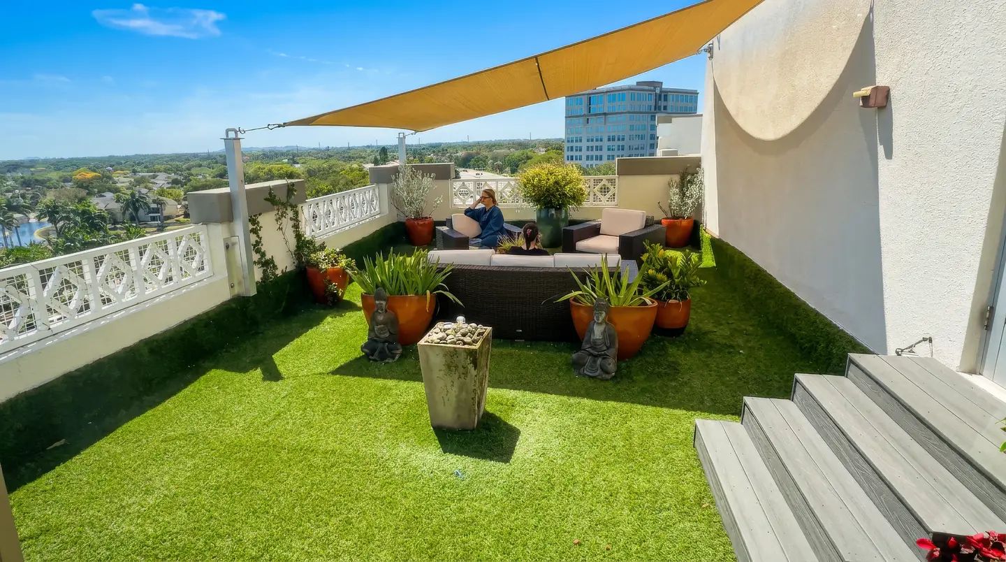 Salon Sora rooftop terrace in Boca Raton: turf lawn, lounge sofa, potted plants, gold shade sail and skyline views (AI-enhanced photo)